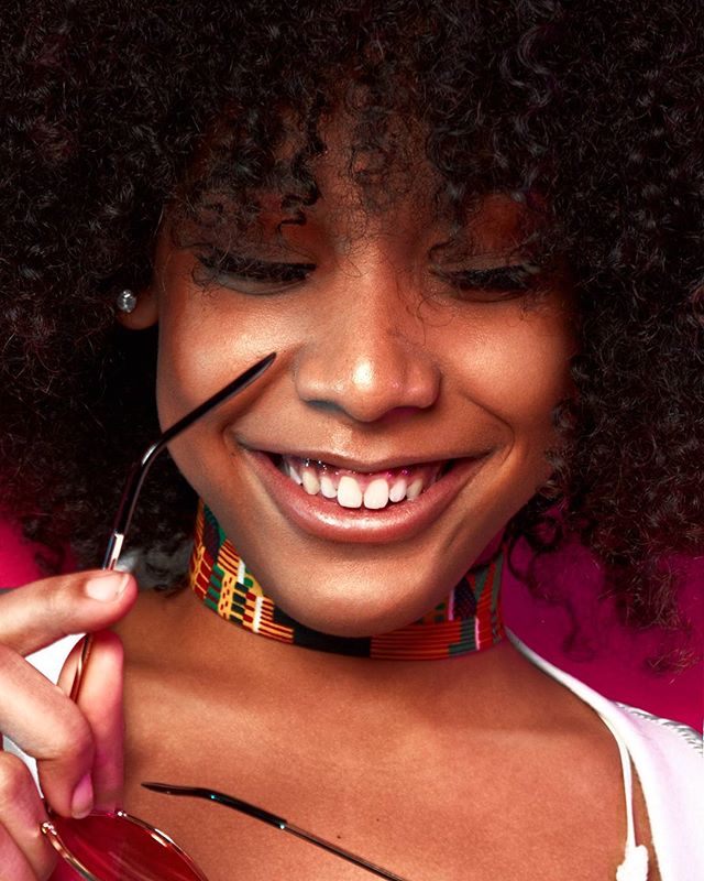 Close-up of a smiling woman with curly hair and a colorful choker holding sunglasses in a studio.
