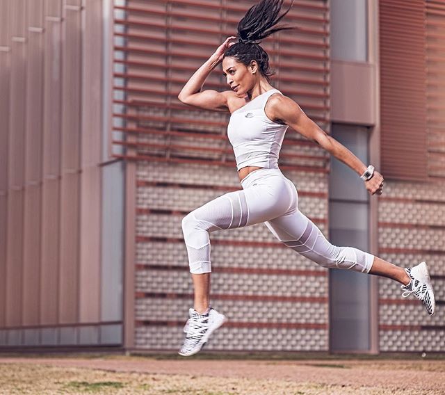 An athletic woman in white sportswear dynamically jumps, conveying energy and strength against an urban backdrop.
