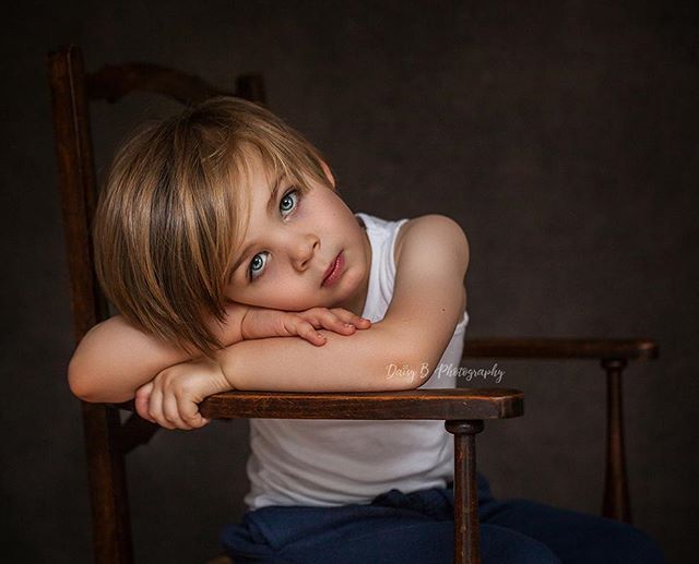 A young boy with blond hair rests his arms on a wooden chair in a softly lit portrait.