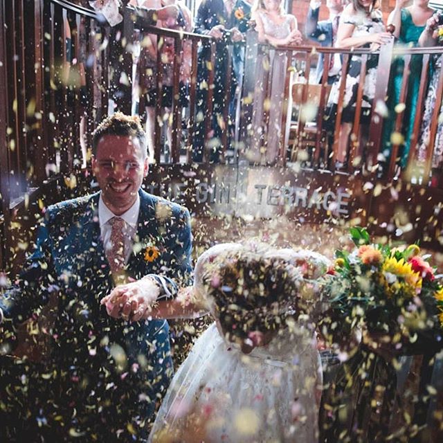 A bride and groom are showered with confetti at their wedding reception, surrounded by happy guests.