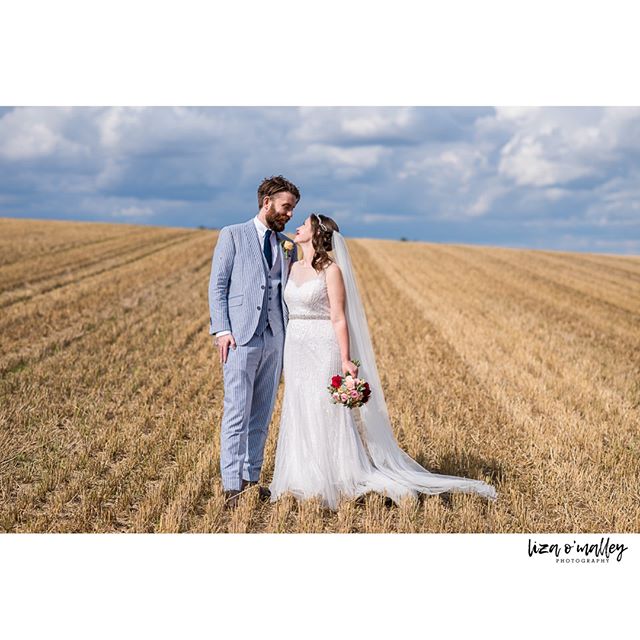 A bride and groom stand lovingly in a golden field on their wedding day under a partly cloudy sky.