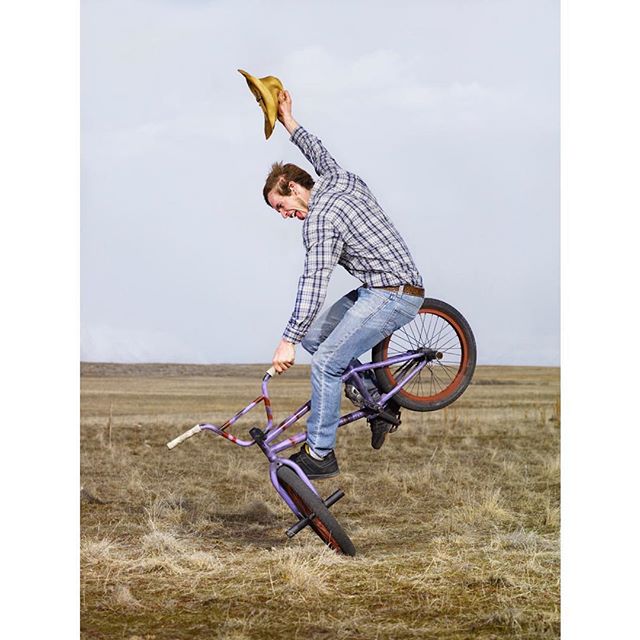 A man performs a wheelie on a BMX bike, tossing his cowboy hat in the air in a field.