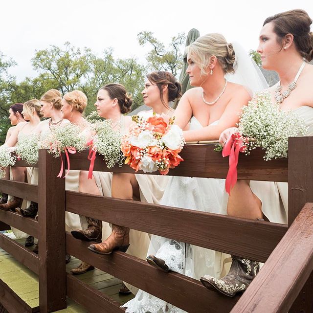 Bridesmaids in cowboy boots lean on a wooden fence, holding bouquets at a country wedding.