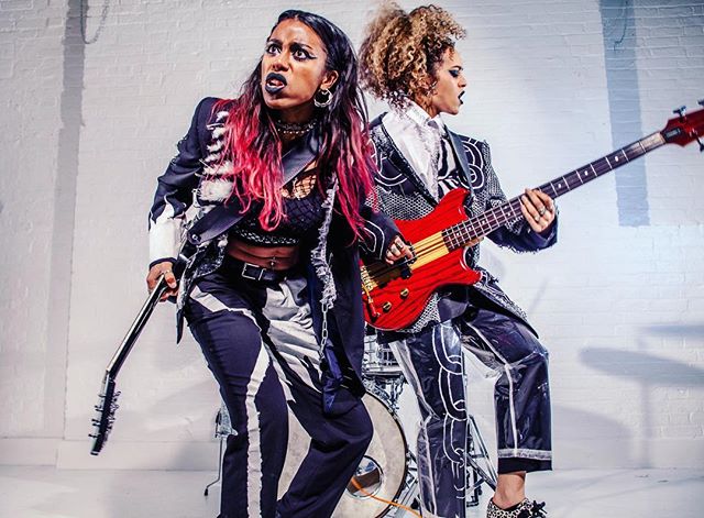 Two female musicians with guitars perform with energy, wearing edgy outfits, in a studio setting.