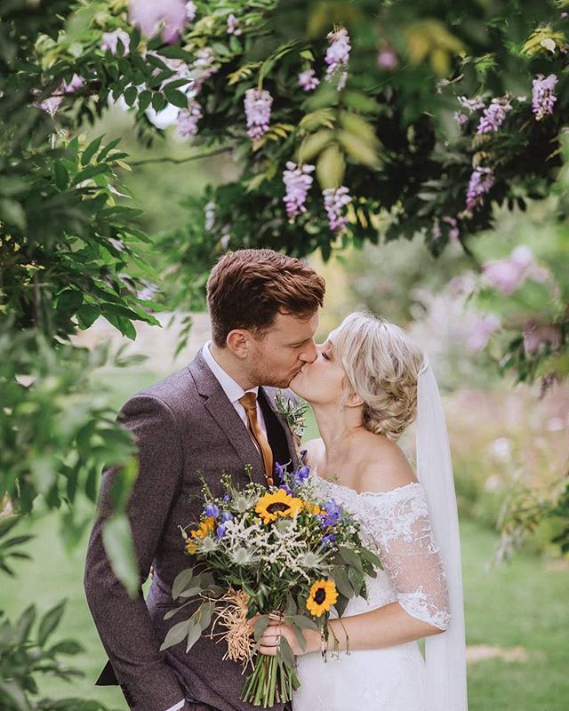 A bride and groom share a kiss surrounded by lush greenery and a colorful bouquet on their wedding day.