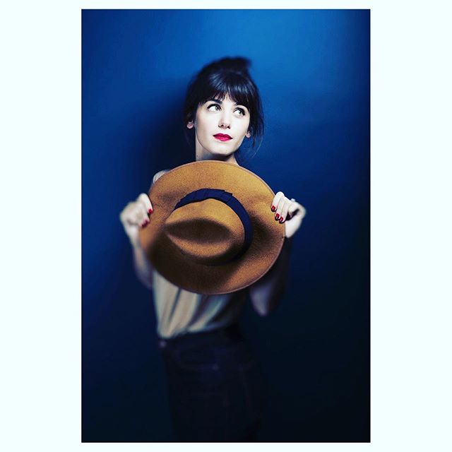 A brunette woman holds a stylish brown hat against a blue backdrop, looking up thoughtfully.