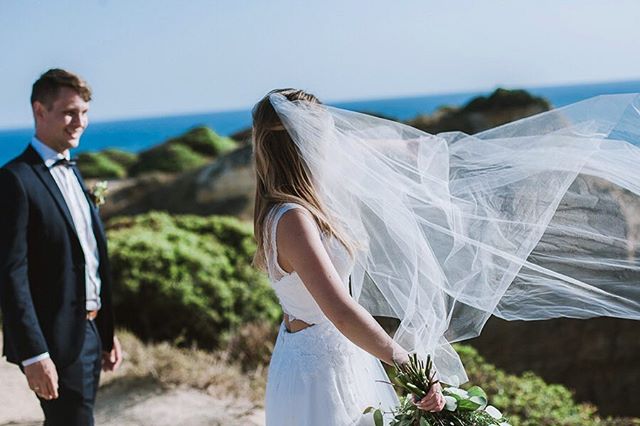 A bride in a veil and a groom are standing outdoors by the coast on their wedding day.
