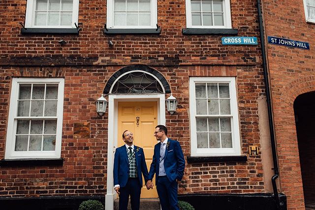 A cheerful same-sex couple celebrates their wedding in front of a brick building on a charming street.