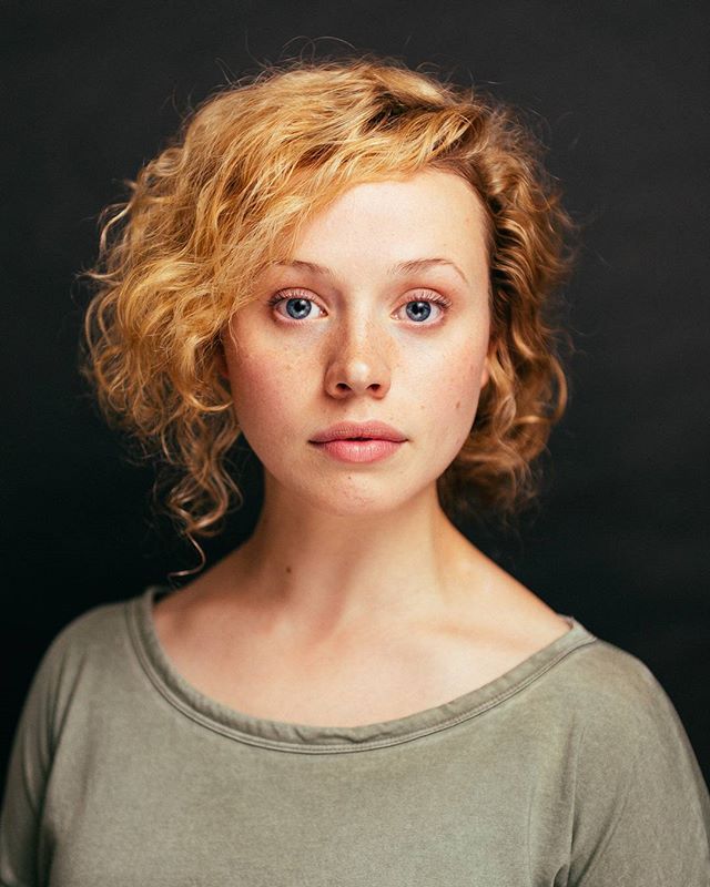A headshot portrait of a young woman with blonde curly hair and blue eyes, wearing a green t-shirt against a dark background.