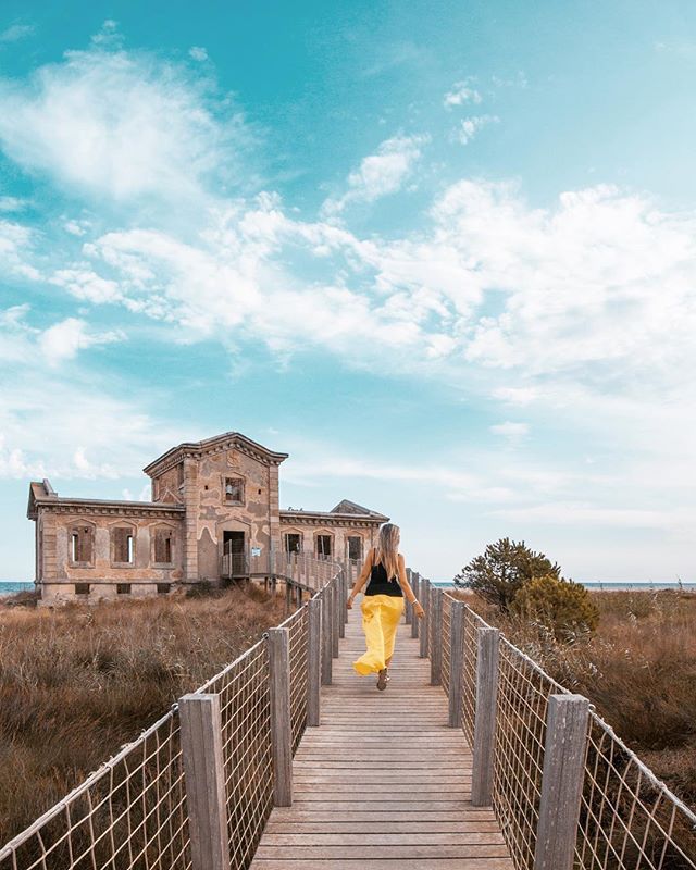 A woman walks on a boardwalk leading to a weathered building under a serene sky.