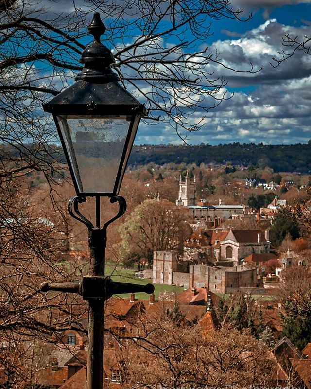 A vintage street light stands overlooking a quaint town with a church spire under a cloudy sky.
