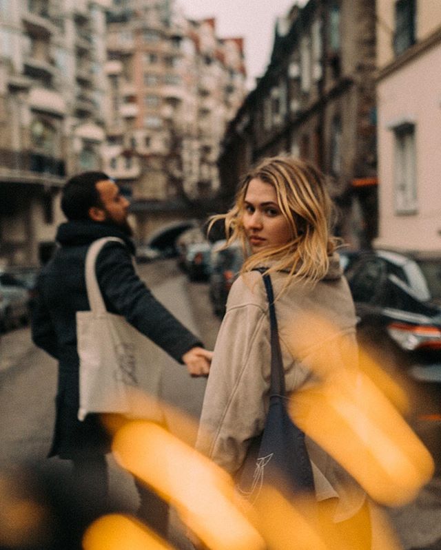 A couple walks hand-in-hand down a city street, the woman glances back at the camera.