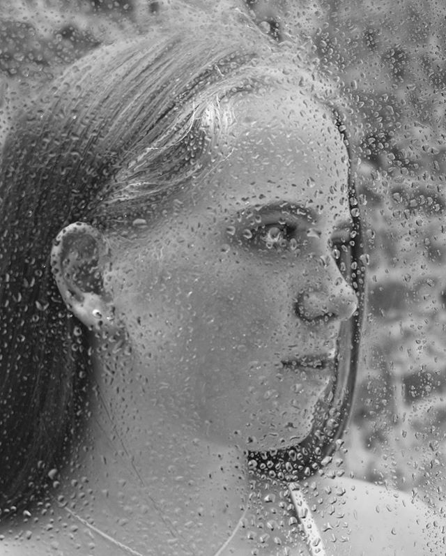 A monochrome portrait of a woman's face seen through a rain-covered glass window, creating a dreamy and emotional atmosphere.