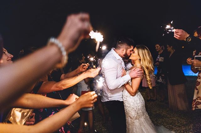 A bride and groom kissing surrounded by wedding guests holding sparklers at a nighttime celebration.