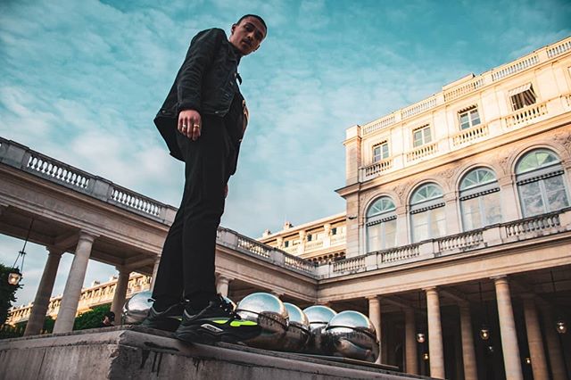 A man in black stands on a wall in front of a building, captured in a low-angle shot.