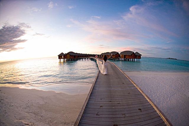 A woman in a white dress walks along a wooden jetty towards bungalows on a tropical beach at sunset.