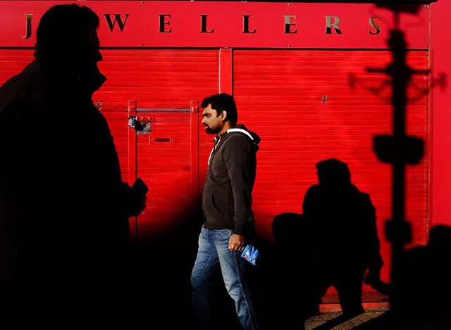 A man walks past a red building with a jeweller's shop sign, casting long shadows on the street.