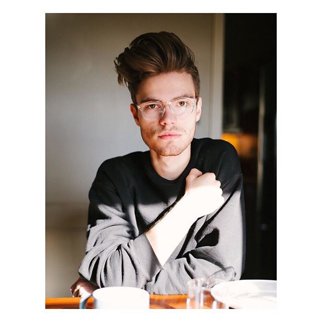 A stylish man with clear eyeglasses sits at a table in a well-lit room, captured in a contemplative portrait.