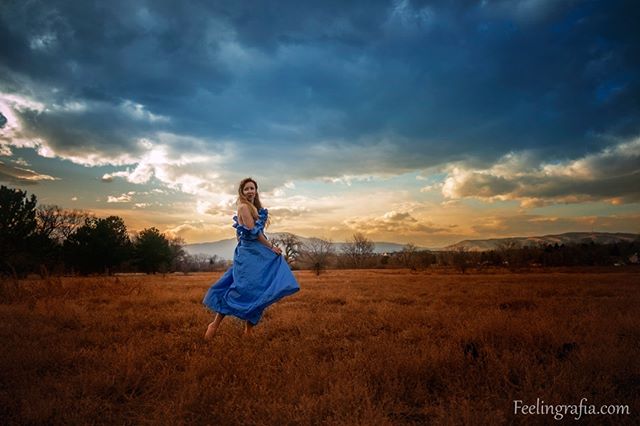 A woman in a blue dress stands in a field with a dramatic sky at sunset.