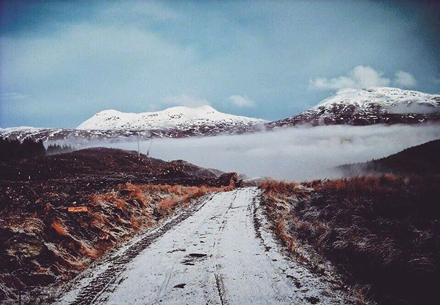 A snowy road winds through a tranquil, mountainous landscape with fog adding to the serene, wintery ambiance.