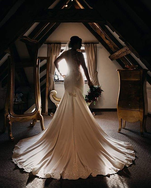 A bride in a wedding dress stands in a room with light from a window behind her.