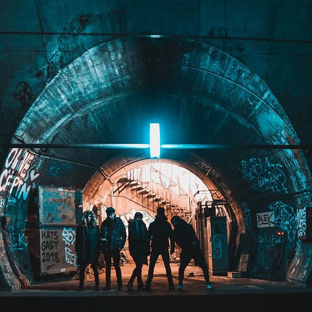 A band poses in an urban tunnel with graffiti, illuminated by a bright light, for a moody editorial shot.