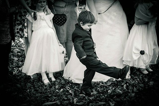 A young boy in a suit playfully kicks leaves at a wedding, surrounded by flower girls and guests.