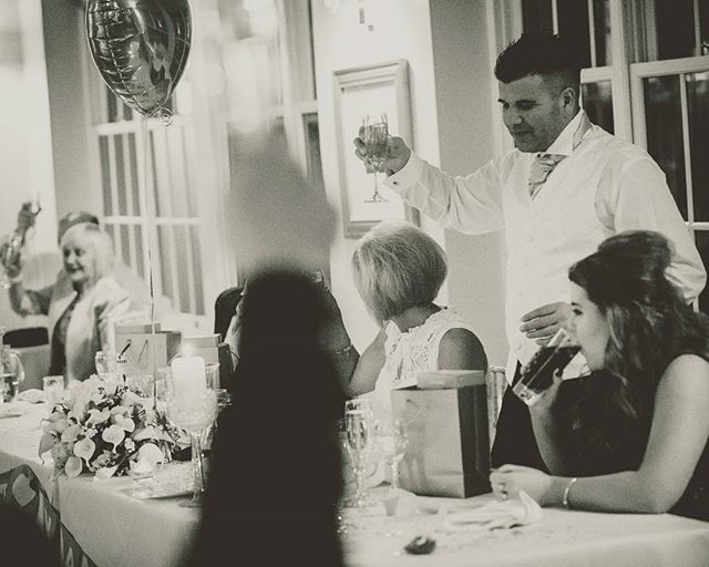 A monochrome image captures a wedding reception with a man giving a toast as guests celebrate.