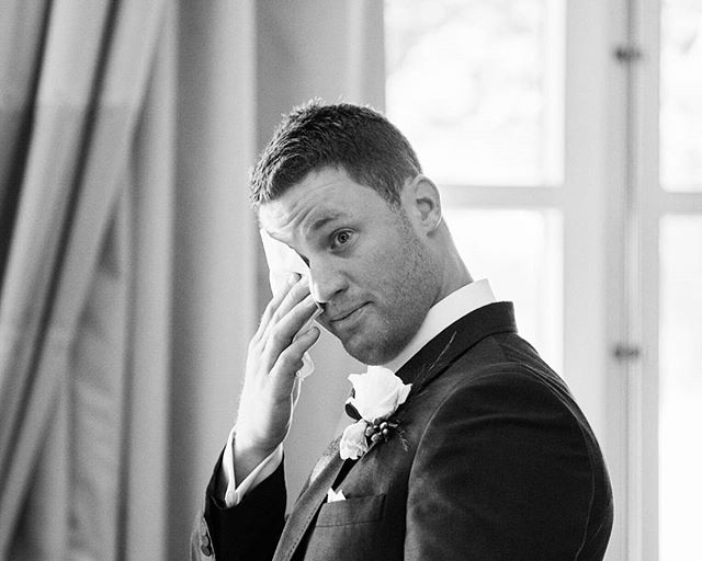 A groom wipes away a tear at his wedding, wearing a suit with a boutonniere.