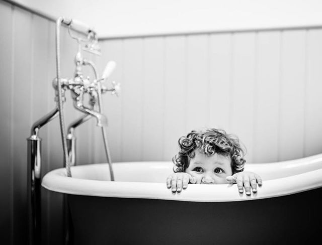 A cute toddler with curly hair peeks over the edge of a vintage bathtub in a playful, black and white photo.