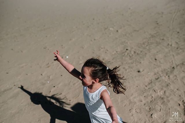 A happy young girl in a white dress reaches up towards the sun while playing on a sandy beach.