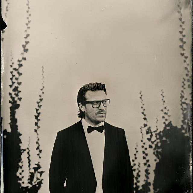 Elegant man in a suit and bow tie wearing glasses, posing in a vintage style black and white photograph.