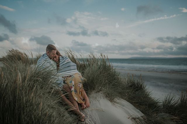 A couple cuddles on a sandy beach dune, wrapped in a blanket enjoying the tranquil ocean view.
