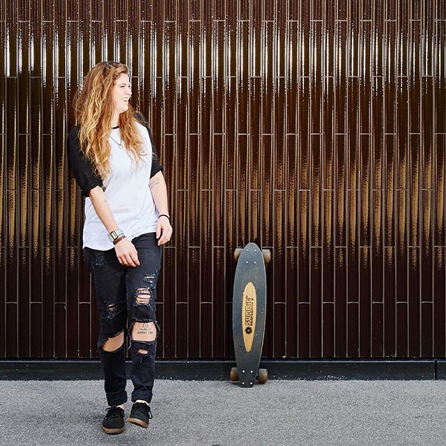 A young woman with a skateboard stands against a textured wall, smiling and wearing ripped jeans.