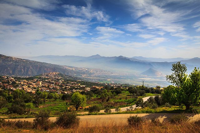 A scenic view of a town nestled in a valley surrounded by hills and mountains under a blue, cloudy sky.