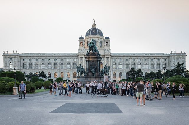 A crowd gathers in front of a historic building and statue in an urban square, likely sightseeing or on a tour.