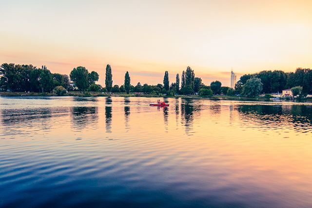 A scenic view of a calm lake at sunset, with people in a boat and trees on the horizon.