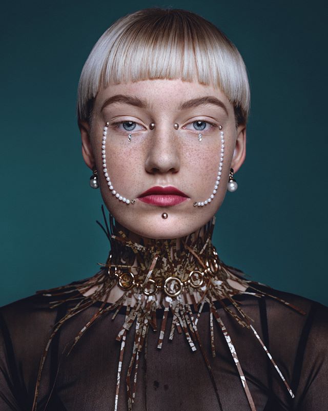 Portrait of a woman with creative makeup, pearl jewelry and short hair, against a teal background.