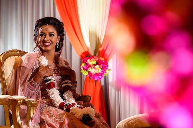 A smiling Indian bride in wedding attire sits gracefully amidst colorful decor for an elegant indoor wedding celebration.