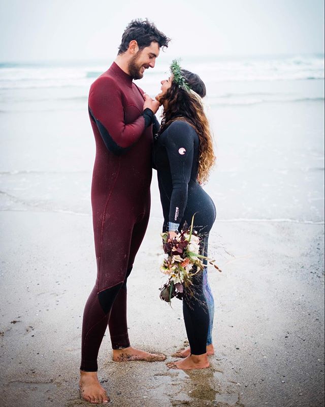 A couple in wetsuits shares an intimate moment on the beach, with the ocean and sky in the background.