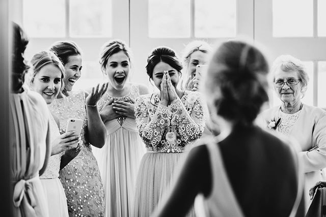 A bride is revealed to her bridesmaids and grandmother, who react with joy at a wedding.