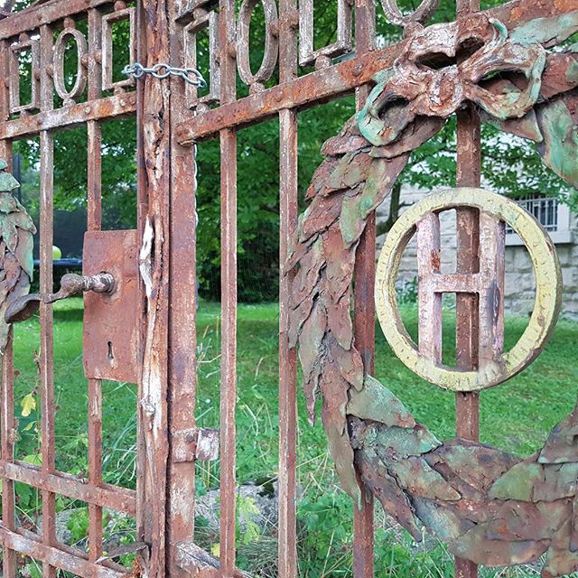 An old, ornate, rusty metal gate with a wreath and the letter H, showing signs of age and history.