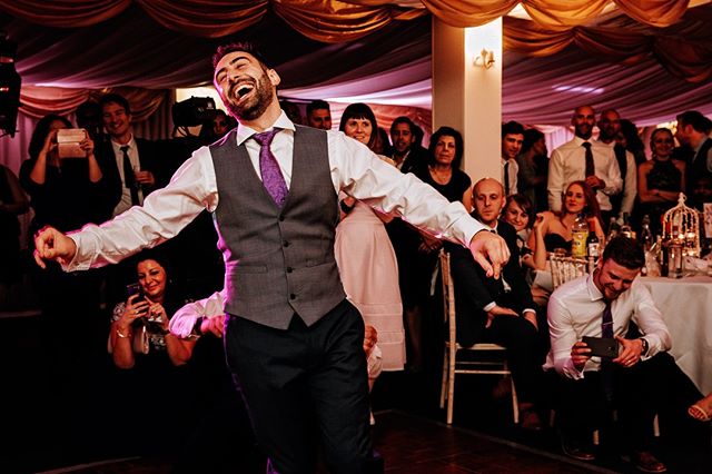 A joyful man dances at a wedding reception, surrounded by celebrating guests in a warmly lit indoor setting.