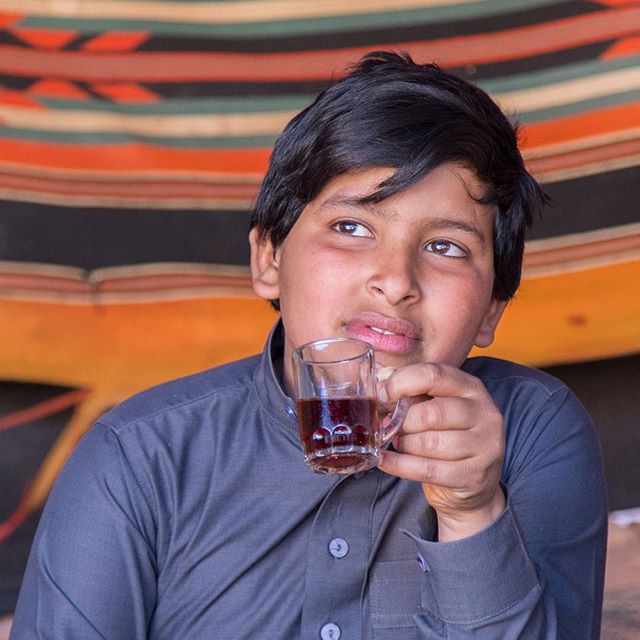 A young boy thoughtfully holds a glass cup of tea, looking away in a cozy indoor setting.