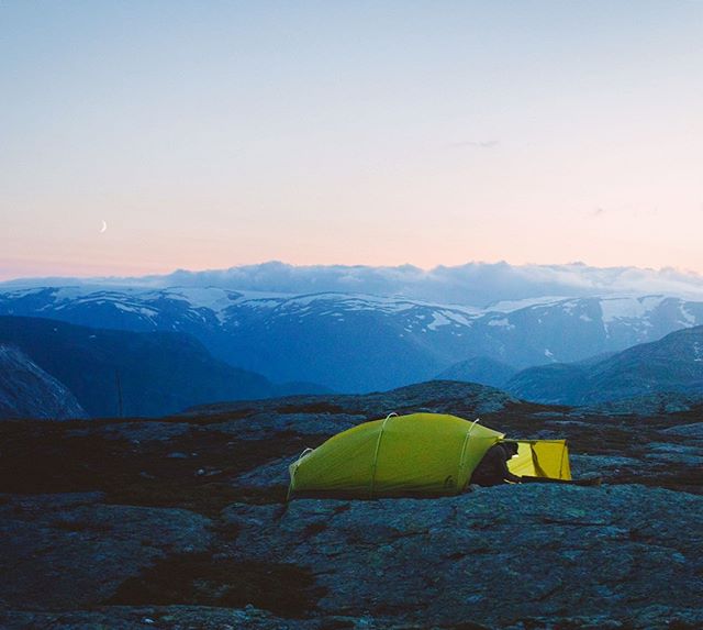 A yellow tent sits on a rocky outcrop against a backdrop of mountains at dusk, evoking a sense of calm and adventure.