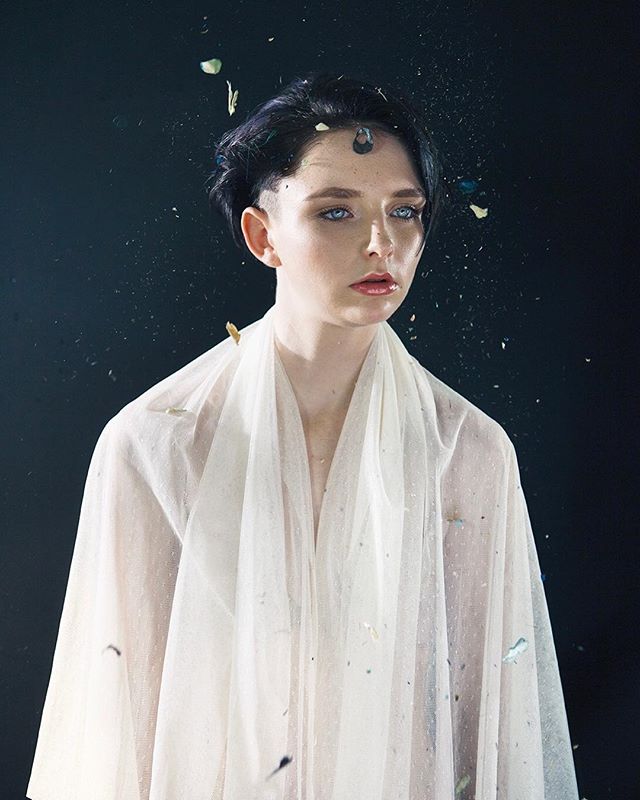 A young woman with blue eyes and short dark hair poses in a sheer white fabric against a dark background.