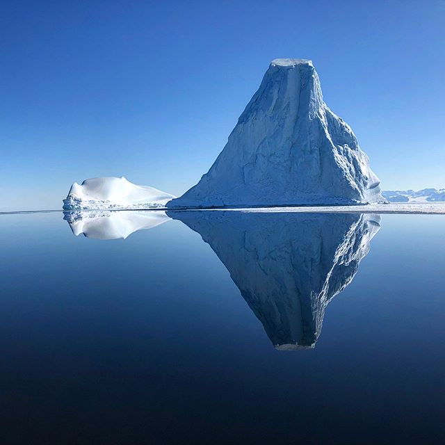 A large iceberg reflects in the clear blue waters of Antarctica under a cloudless sky.