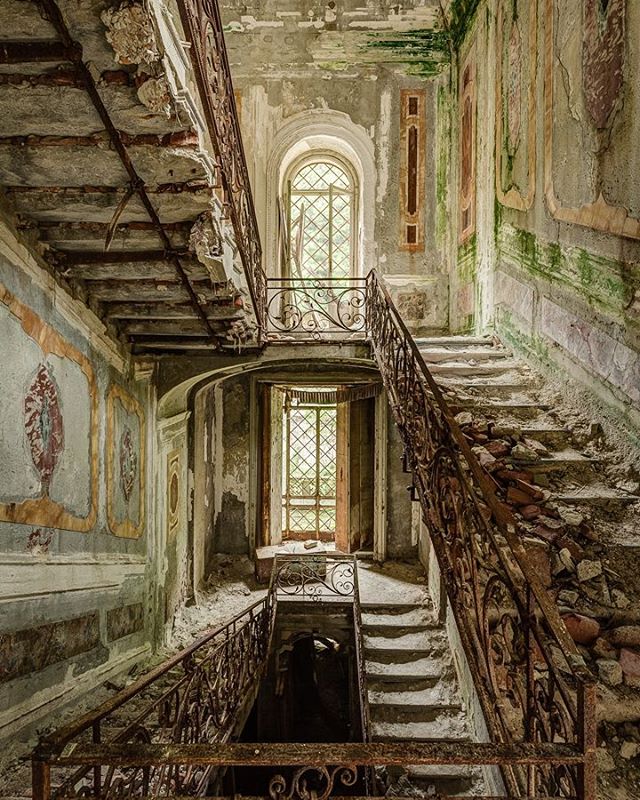 An abandoned interior features a staircase and ornate design details, creating a vintage and elegant atmosphere.