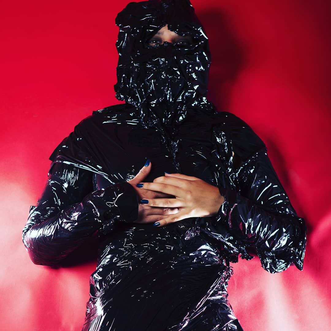 A person wrapped in shiny black plastic against a red background, creating a mysterious and avant-garde editorial image.