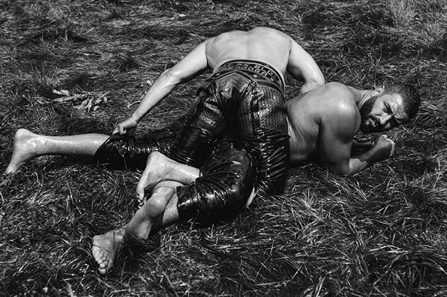 Two shirtless oil wrestlers grapple on a grassy field in a black and white, medium shot.